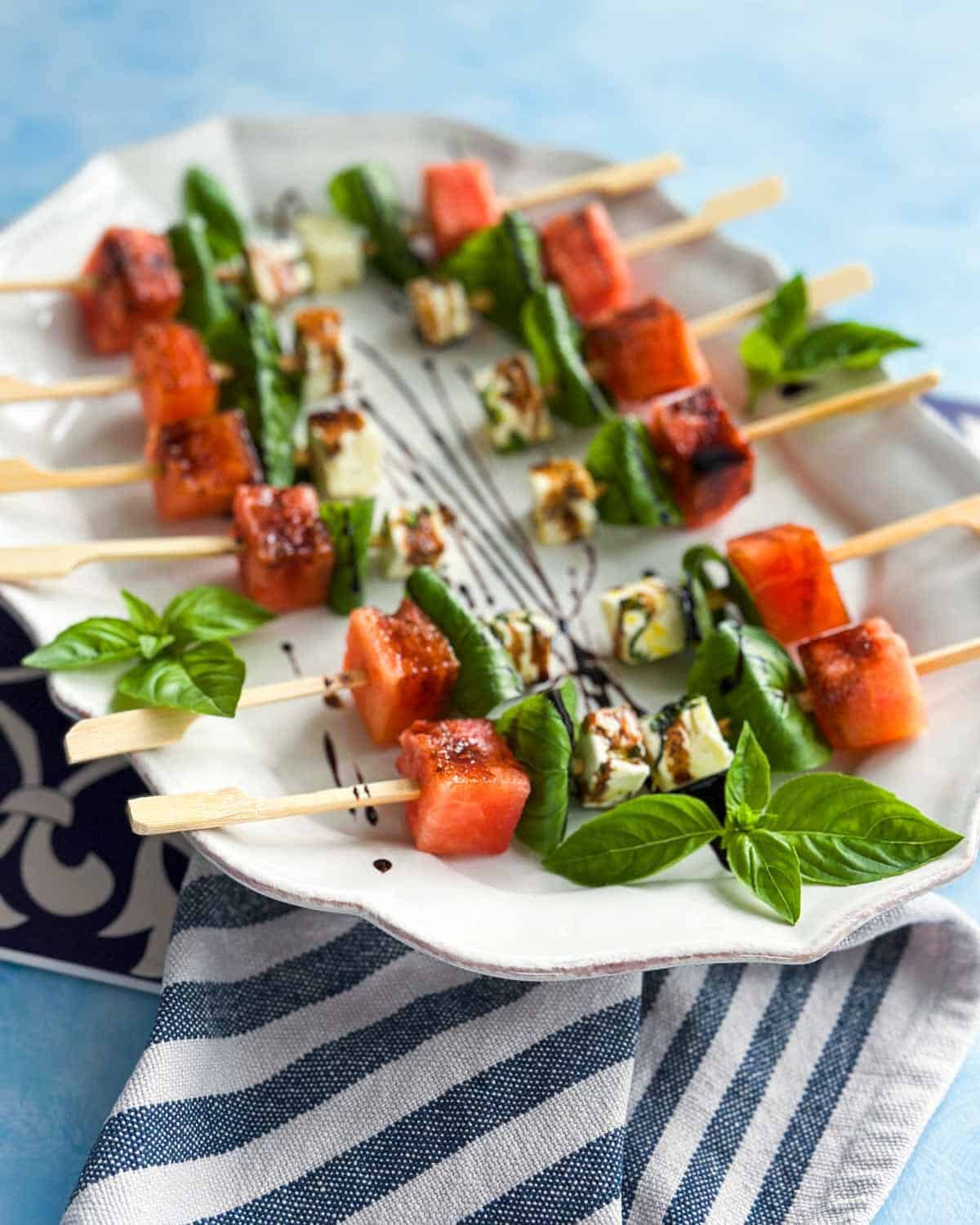 Top-down view of watermelon feta skewers arranged on a white serving platter with fresh basil leaves and balsamic glaze, placed over a striped cloth with a soft blue background.