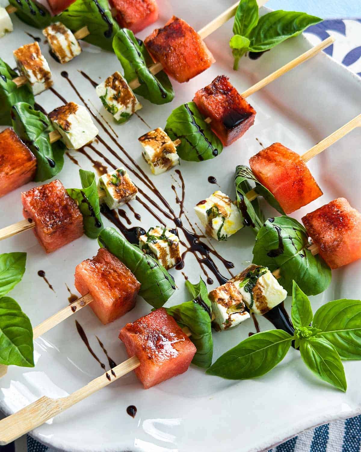 Close-up view of watermelon feta skewers arranged neatly on a white serving plate, highlighting the contrast of sweet watermelon, herby feta cubes, and vibrant basil leaves with balsamic glaze.