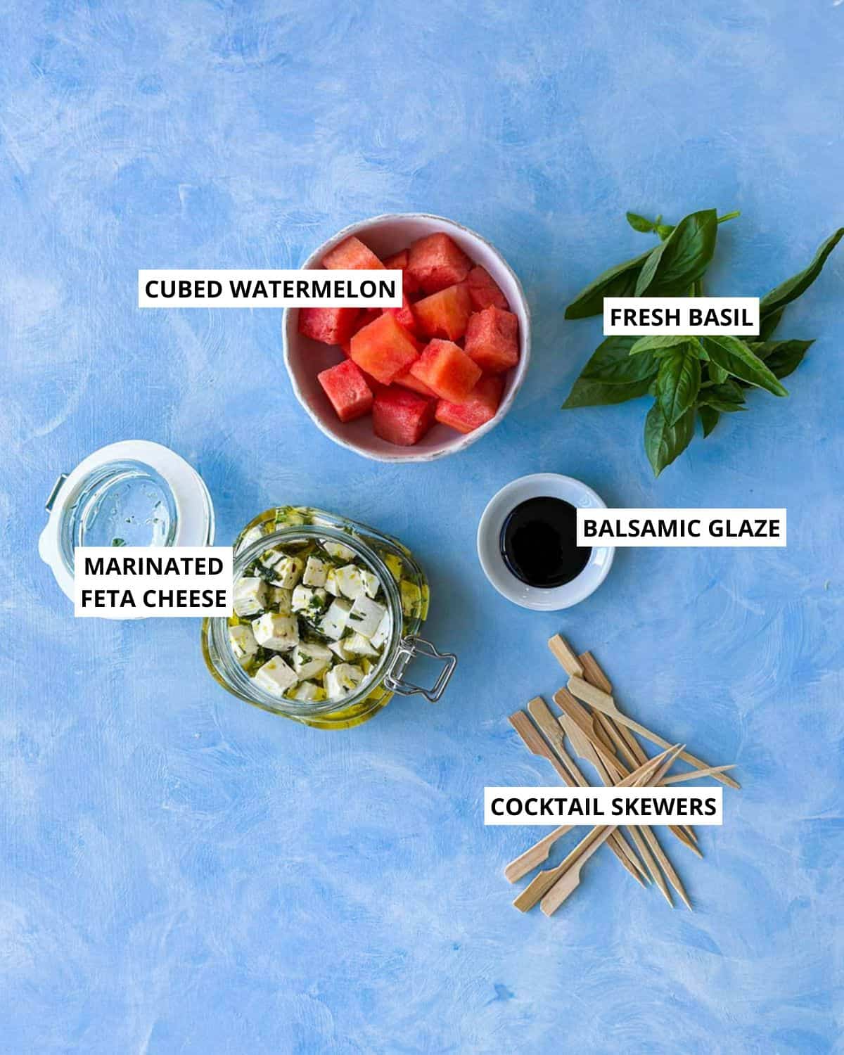 op-down view of ingredients for watermelon feta skewers on a blue background, including cubed watermelon, marinated feta cheese, fresh basil, balsamic glaze, and cocktail skewers, each clearly labeled.