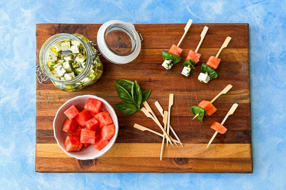 Overhead view of a wooden board with cubed watermelon, marinated feta, fresh basil leaves, and cocktail skewers, showing the step-by-step assembly of watermelon feta skewers.