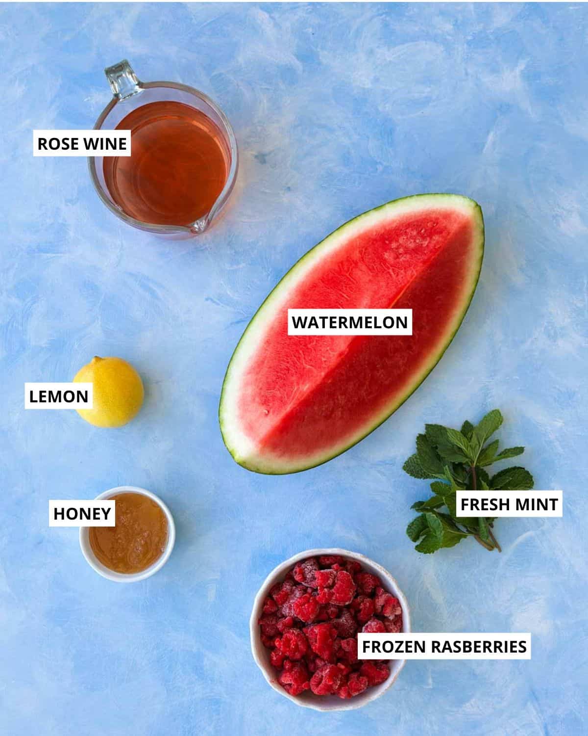 Overhead flat lay of watermelon frosé ingredients including rosé wine, watermelon, frozen raspberries, lemon, honey, and fresh mint on a pale blue background.