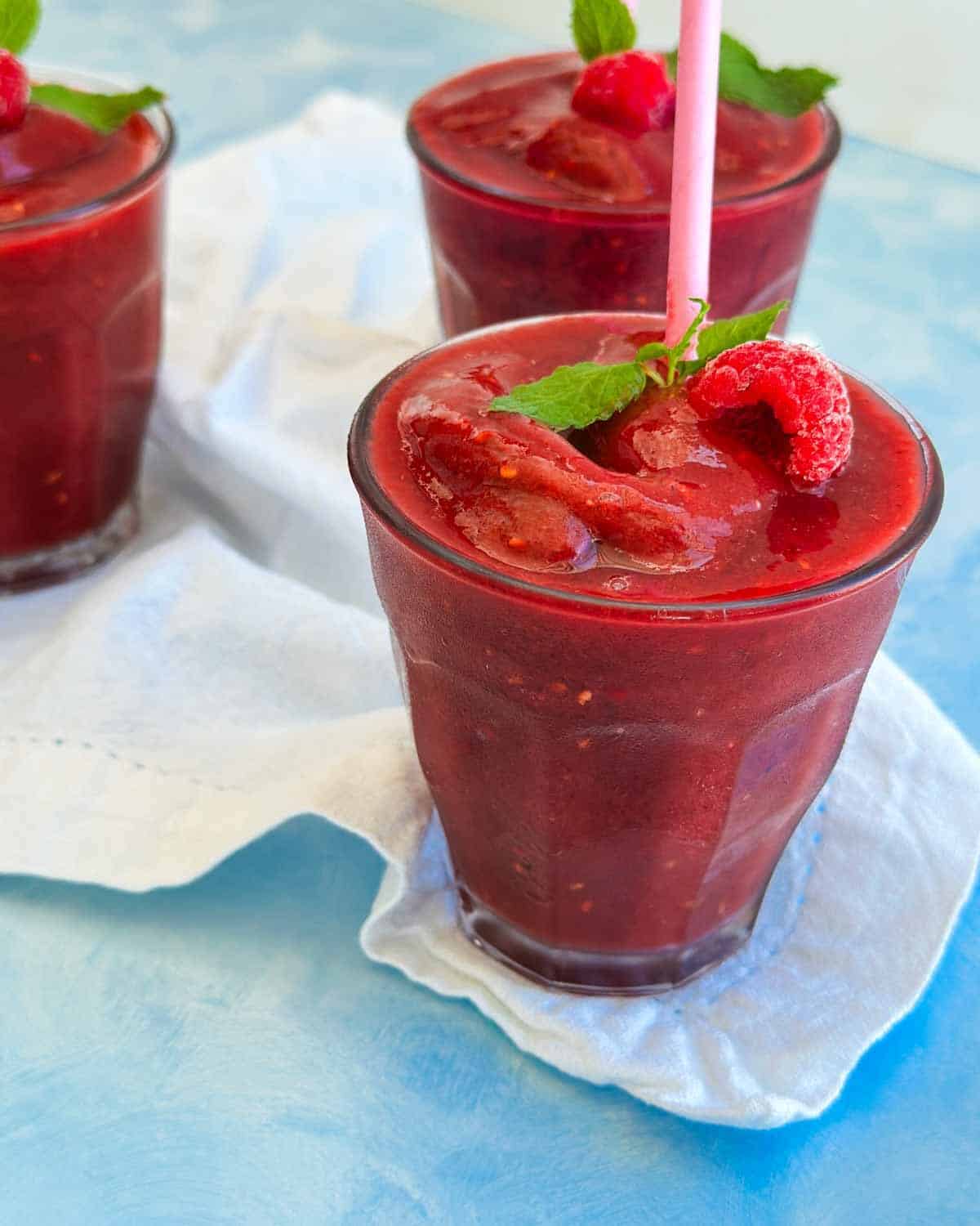 Close-up watermelon slushie with berries showing its smooth frozen texture, raspberry garnish, mint leaves, and pink paper straw.