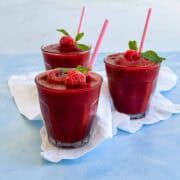 Watermelon slushie garnished with frozen raspberries, mint leaves, and pink paper straws, served in clear glasses on a light blue tabletop.