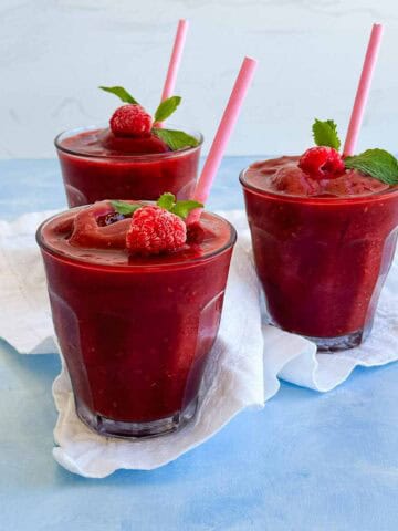 Watermelon slushie garnished with frozen raspberries, mint leaves, and pink paper straws, served in clear glasses on a light blue tabletop.