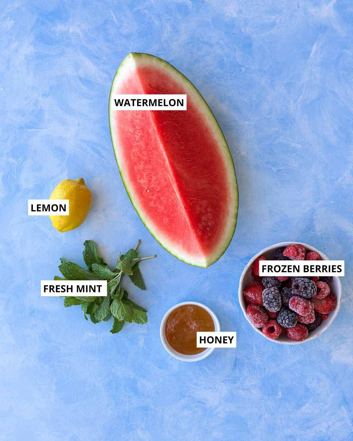 Ingredients for watermelon slushie including fresh watermelon, frozen mixed berries, lemon, honey, and fresh mint arranged on a blue surface.