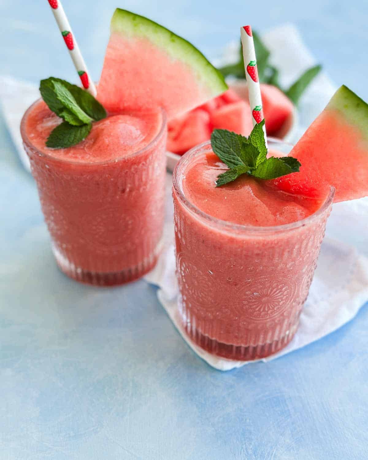 Two watermelon smoothies served in glasses with mint, watermelon wedges, and a bowl of fresh watermelon cubes.