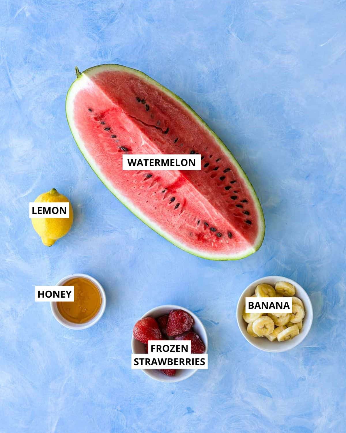 Top-down view of watermelon smoothie ingredients with watermelon, lemon, honey, frozen strawberries, and banana.
