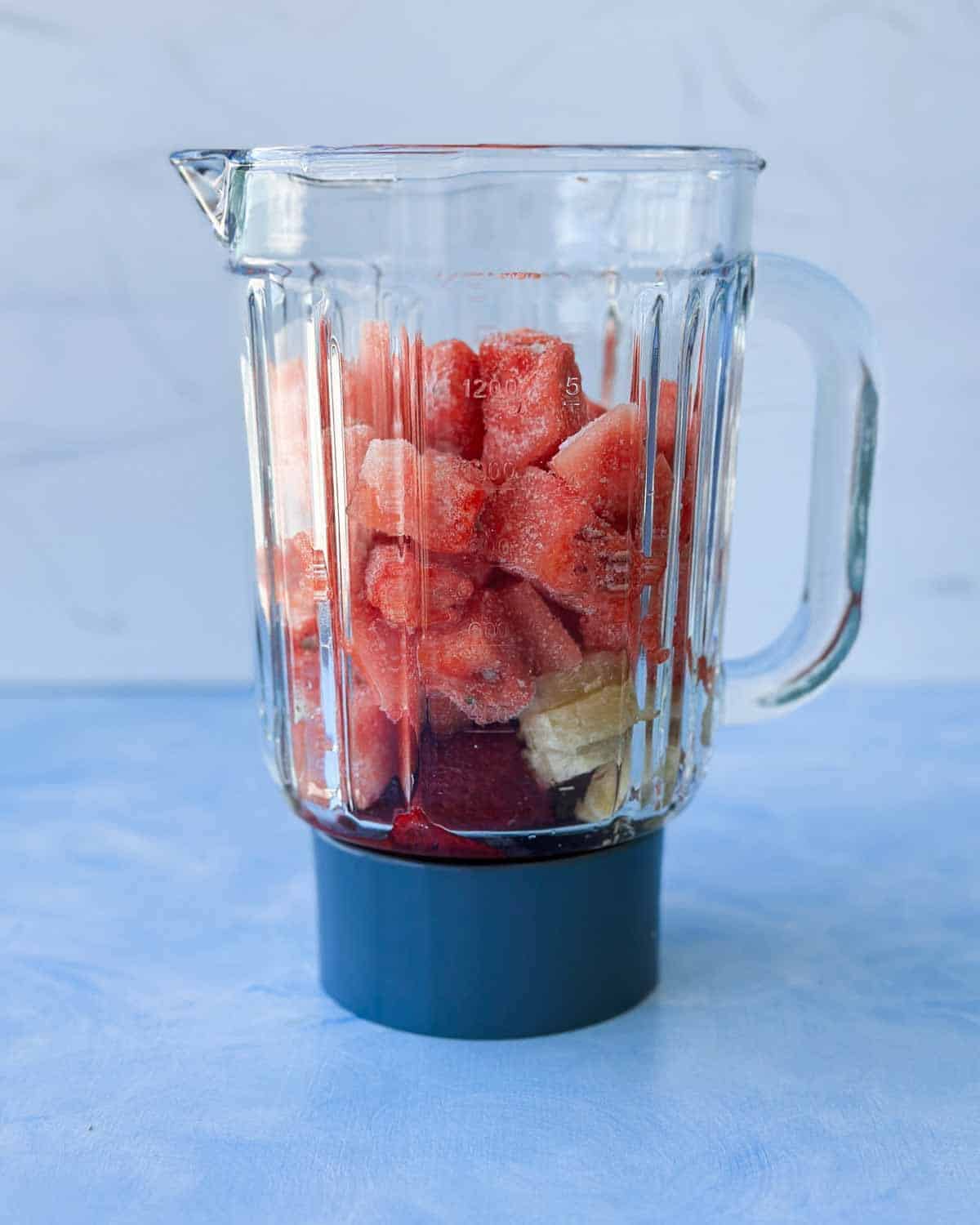 Watermelon, frozen strawberries, and banana added to a glass blender jug before blending.