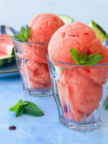 Watermelon sorbet scooped into glass dessert cups with fresh mint leaves and watermelon wedges, styled on a light blue surface with sliced watermelon behind.