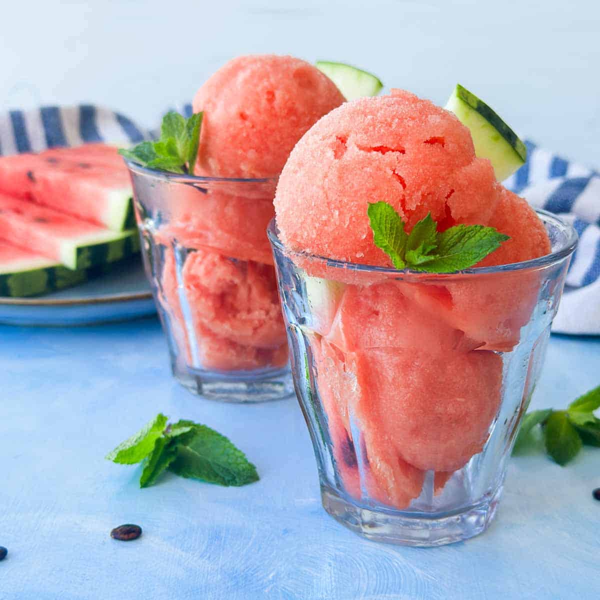 Watermelon sorbet scooped into glass dessert cups with fresh mint leaves and watermelon wedges, styled on a light blue surface with sliced watermelon behind.
