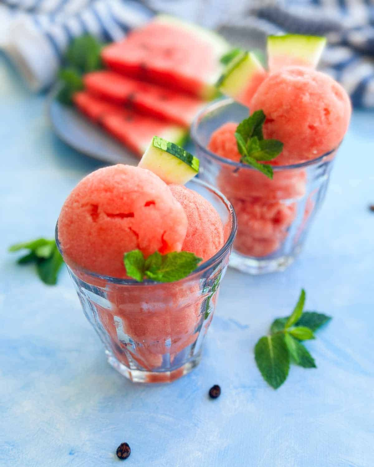 Watermelon sorbet scoops served in small glass cups, garnished with fresh mint and watermelon wedges, photographed with fresh watermelon slices in the background.