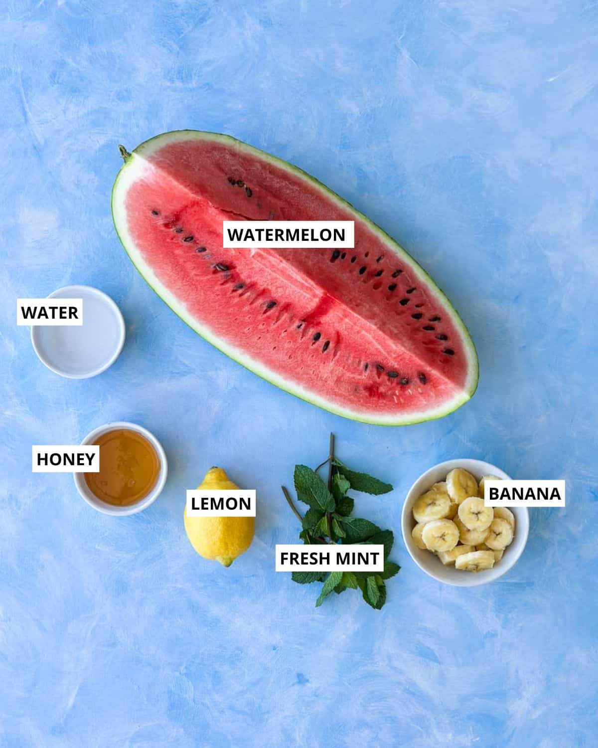 Overhead view of ingredients for watermelon sorbet including watermelon, banana, honey, lemon, fresh mint, and water arranged on a light blue surface.