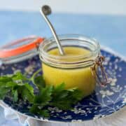 Close-up of homemade white wine vinaigrette in a small glass jar with a spoon, resting on a blue floral plate with fresh parsley.