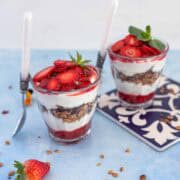 Two strawberry yogurt parfaits with layers of yogurt, granola, and fresh strawberries, served in glasses with spoons and styled on a decorative tile.