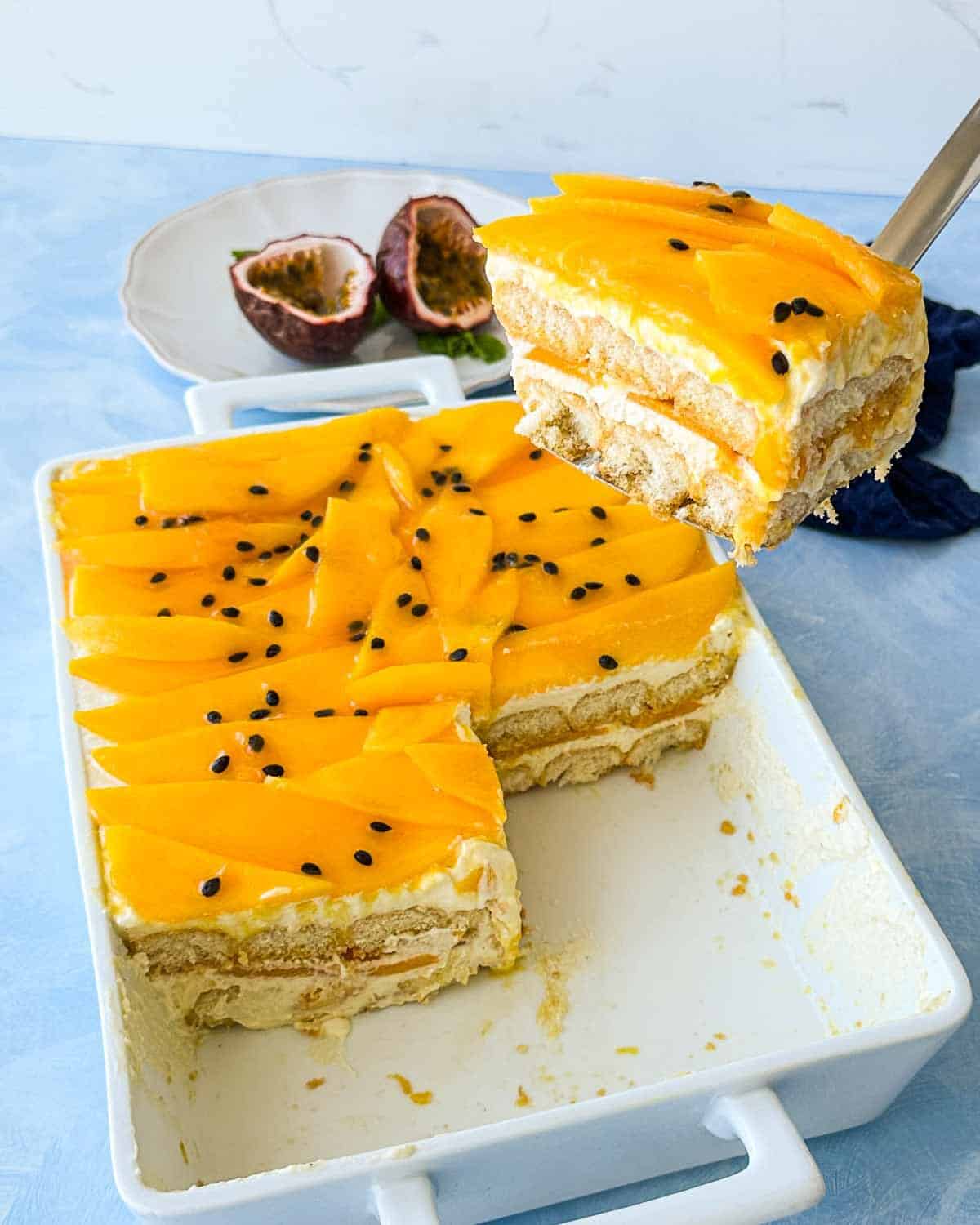 Angled view of a large slice of mango tiramisu being lifted from a white baking dish, revealing distinct layers of cream, mango, and ladyfingers, with fresh mango slices and passion fruit seeds on top and halved passion fruit in the background.