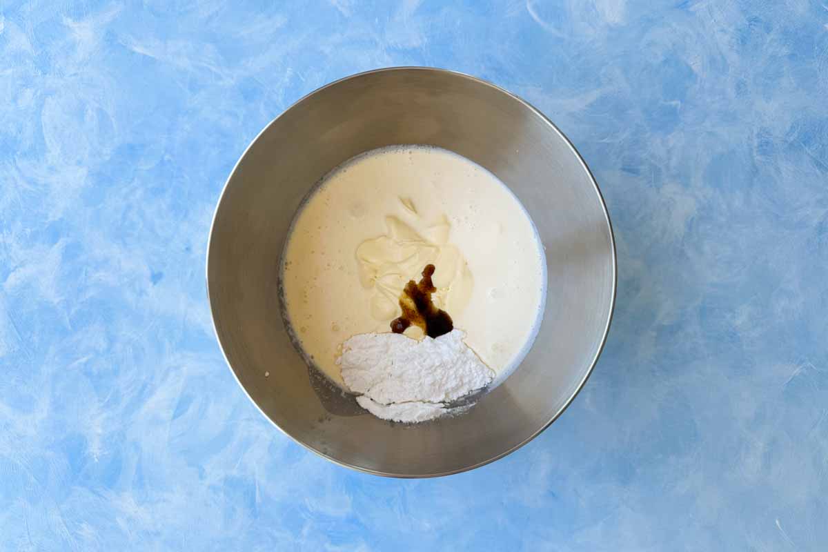 Top view of a mixing bowl with heavy cream, mascarpone, powdered sugar, and vanilla bean paste added together, ready to be whipped into a smooth cream mixture on a light blue background.