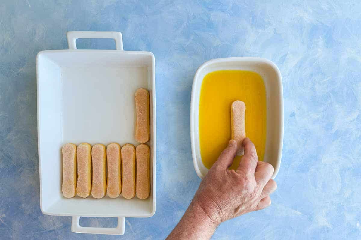 Top view of a hand dipping savoiardi biscuits into orange juice beside a white baking dish lined with soaked biscuits, showing the start of the tiramisu layering process.