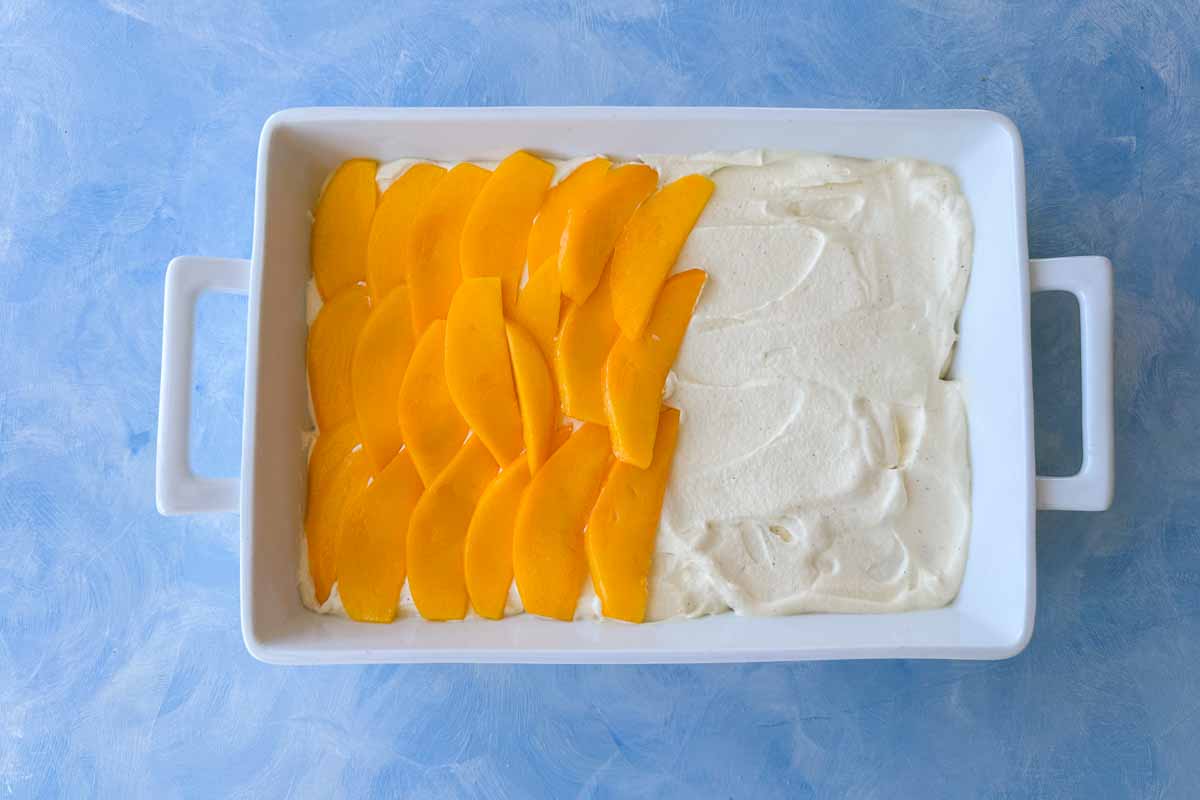 Top view of a white baking dish with a layer of mascarpone cream spread on one side and fresh mango slices arranged on the other, showing the layering step in progress.