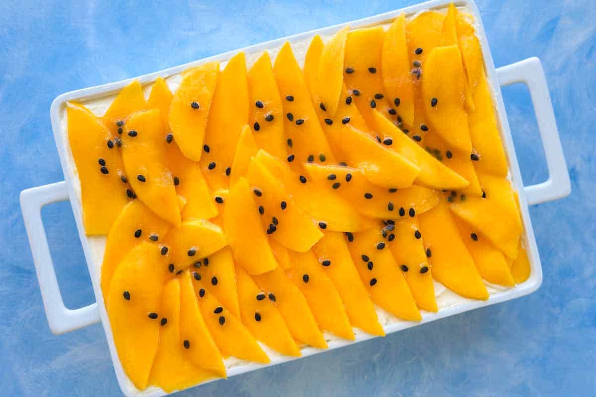 Top view of the finished mango tiramisu topped with glossy mango slices and scattered passion fruit seeds, set in a white baking dish on a light blue textured surface.