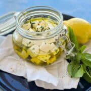 Three-quarter angle view of a glass jar of marinated feta showing layers of creamy feta cubes, olive oil, and herbs, with a lemon and fresh basil and mint placed beside it on a dark plate.