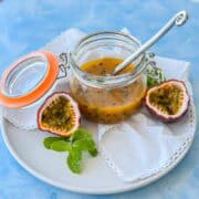 Angled close-up of a spoon resting inside a jar of passion fruit coulis, with fresh passion fruit halves and mint leaves on a white plate, showing the thick, seed-studded texture.