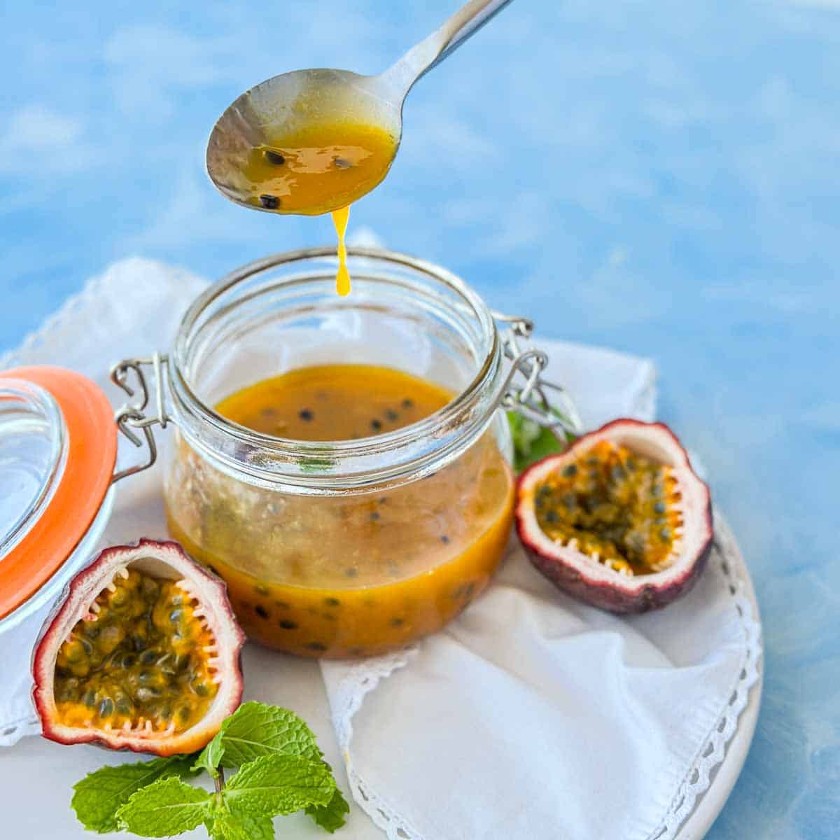Close-up of passion fruit coulis dripping from a spoon back into a glass jar, highlighting the smooth, glossy consistency and speckled seeds, with fresh fruit and mint in the background.