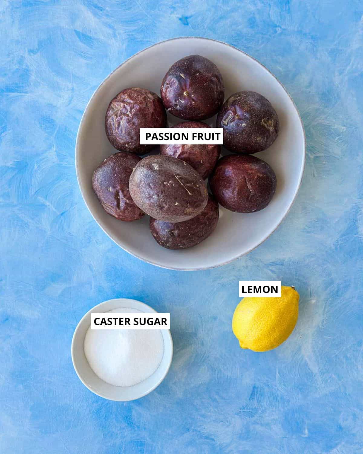 Overhead layout of fresh passion fruits in a bowl, a whole lemon, and a small dish of caster sugar arranged on a light blue textured surface with labeled ingredients.