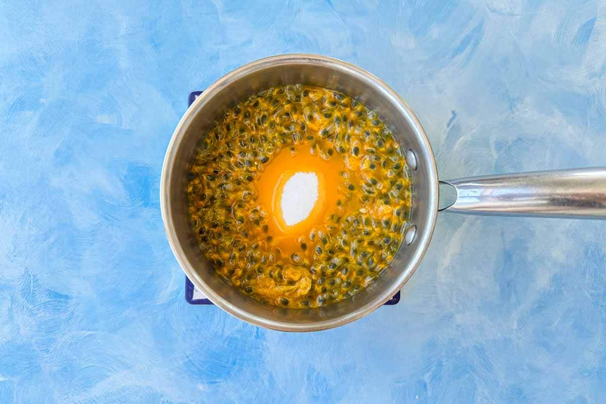 Top-down view of a saucepan filled with fresh passion fruit pulp and caster sugar before cooking, showing the vibrant orange color and seeds against a light blue background.