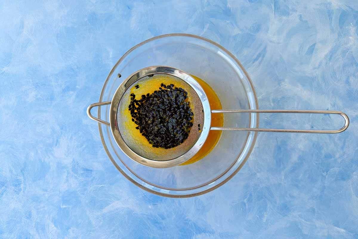 Overhead close-up of strained passion fruit coulis being passed through a fine mesh sieve into a glass bowl, separating the seeds from the smooth golden sauce underneath.