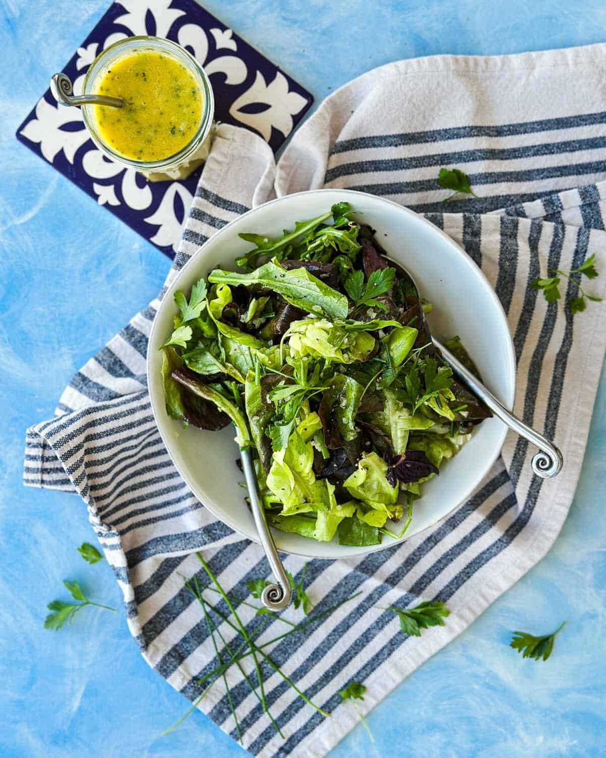 Overhead view of a bistro salad in a white bowl with mixed greens, herbs, and a jar of vinaigrette on the side.