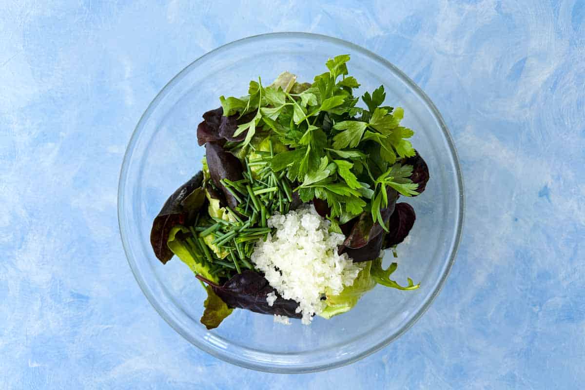 Top view of mixed salad greens, chopped shallots, parsley, and chives added to a glass bowl for bistro salad.