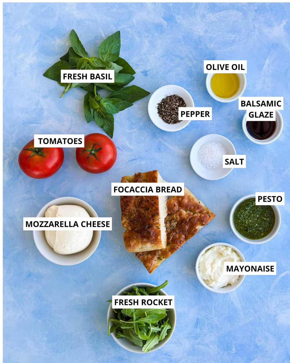 Top-down ingredient layout for Caprese Chicken sandwiches with focaccia, tomatoes, mozzarella, pesto, basil, rocket, and mayo.