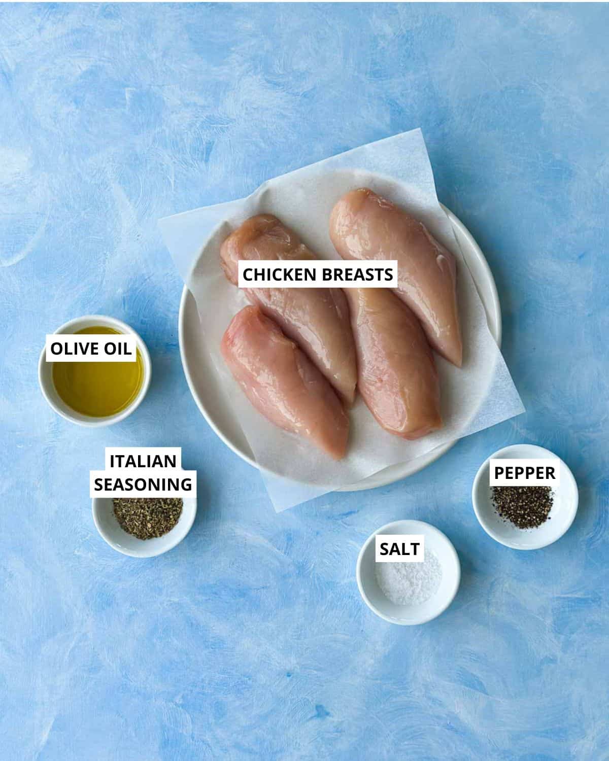 Top-down view of chicken breasts, olive oil, Italian seasoning, salt, and pepper arranged on a blue surface.