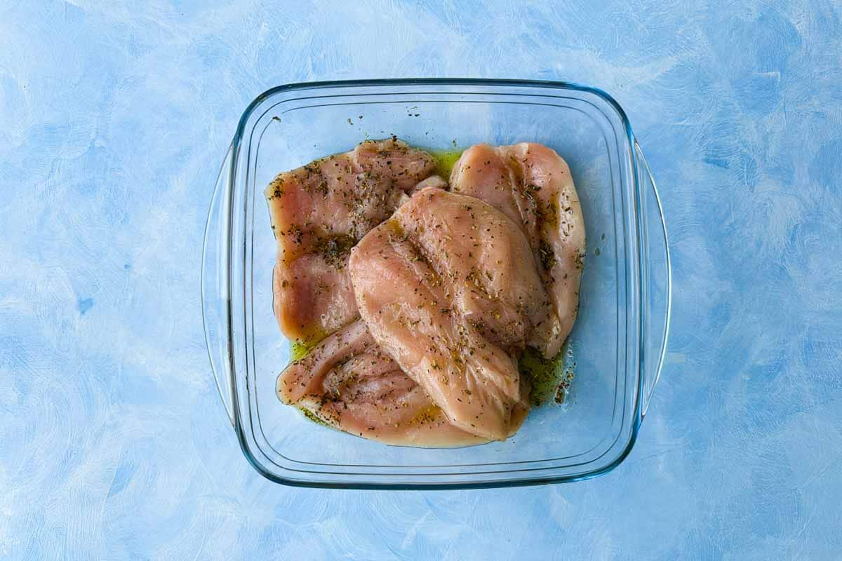 Chicken breasts marinating in olive oil, Italian seasoning, salt, and pepper in a glass dish on a blue surface.