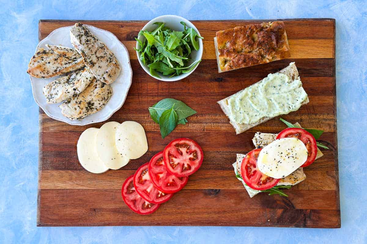 Top-down view of chicken Caprese sandwiches being assembled with focaccia, pesto mayo, chicken, tomato, mozzarella, and rocket.