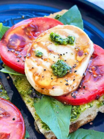 Close-up view of a pesto caprese sandwich with creamy mozzarella, ripe tomato, basil leaves, and glossy balsamic glaze.