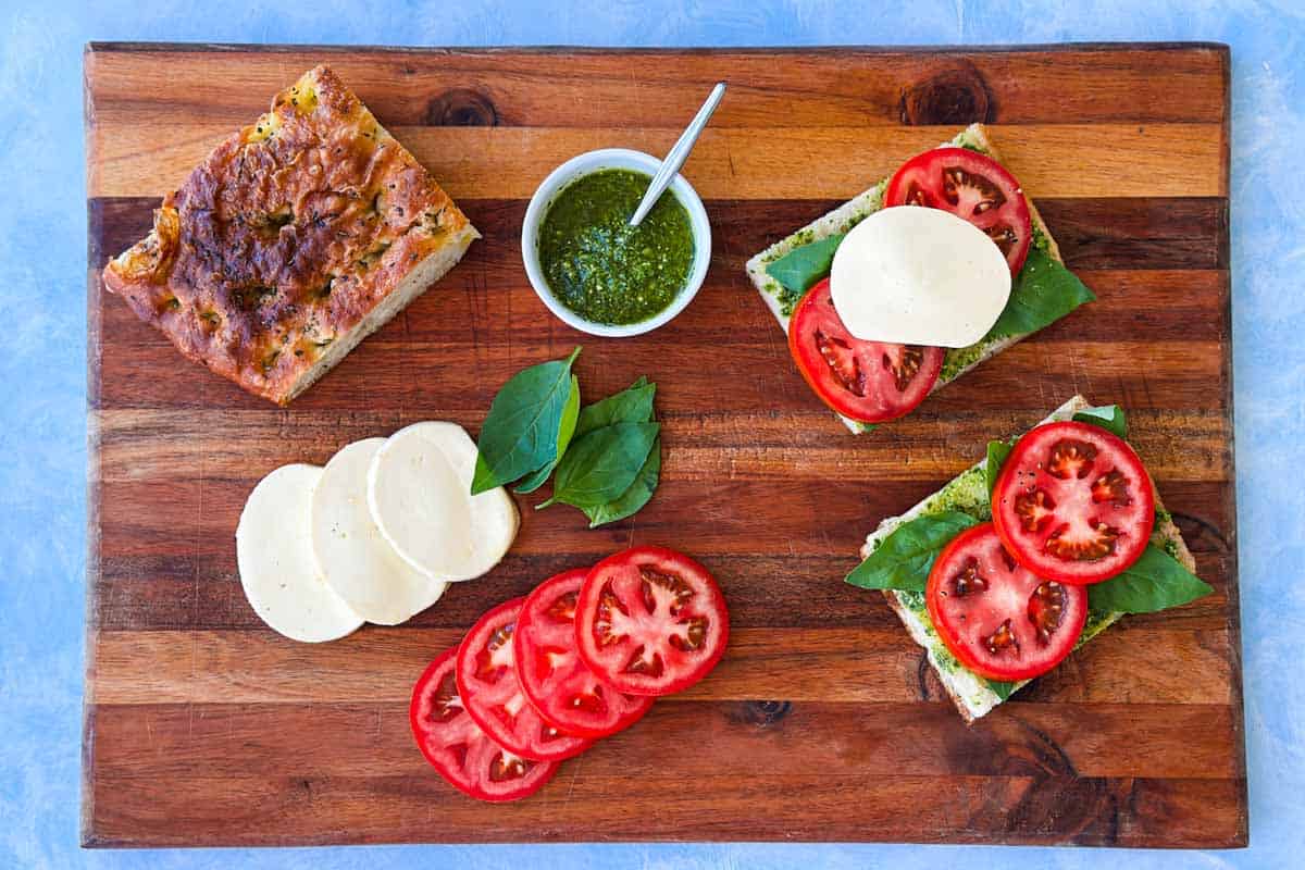 Top view of pesto caprese sandwich ingredients being assembled, on a wooden board, with focaccia, tomato slices, mozzarella, basil, and pesto.