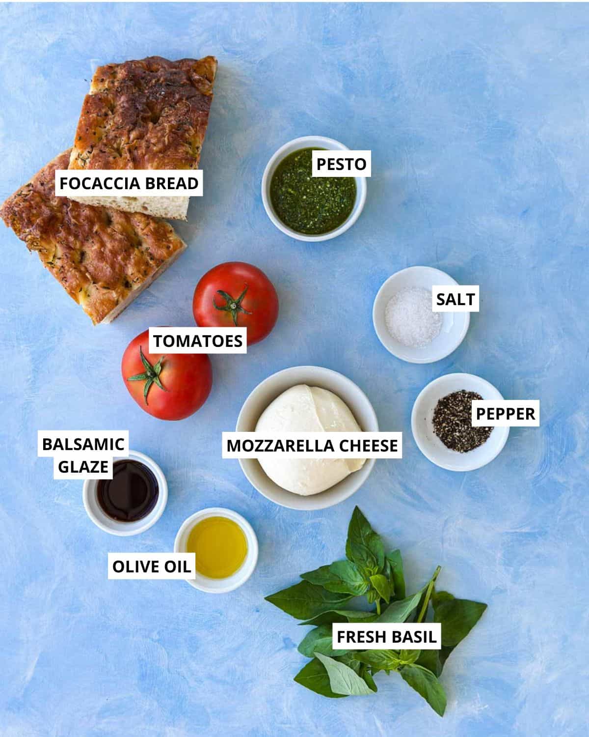 Flat lay of pesto caprese sandwich ingredients, including focaccia, pesto, tomatoes, mozzarella, basil, olive oil, and balsamic glaze.