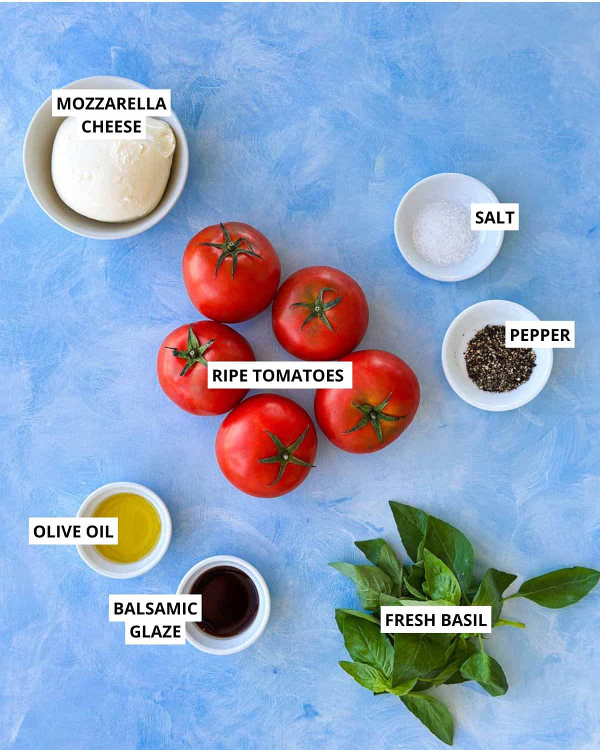 Flat lay of caprese salad ingredients on a blue surface, including whole ripe tomatoes, fresh mozzarella, basil leaves, olive oil, balsamic glaze, salt and pepper arranged in small bowls.