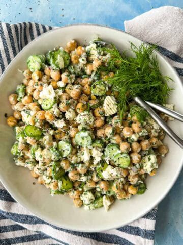 Top-down view of chickpea and feta salad with cucumbers, fresh dill, herbs, and creamy dressing in a serving bowl.