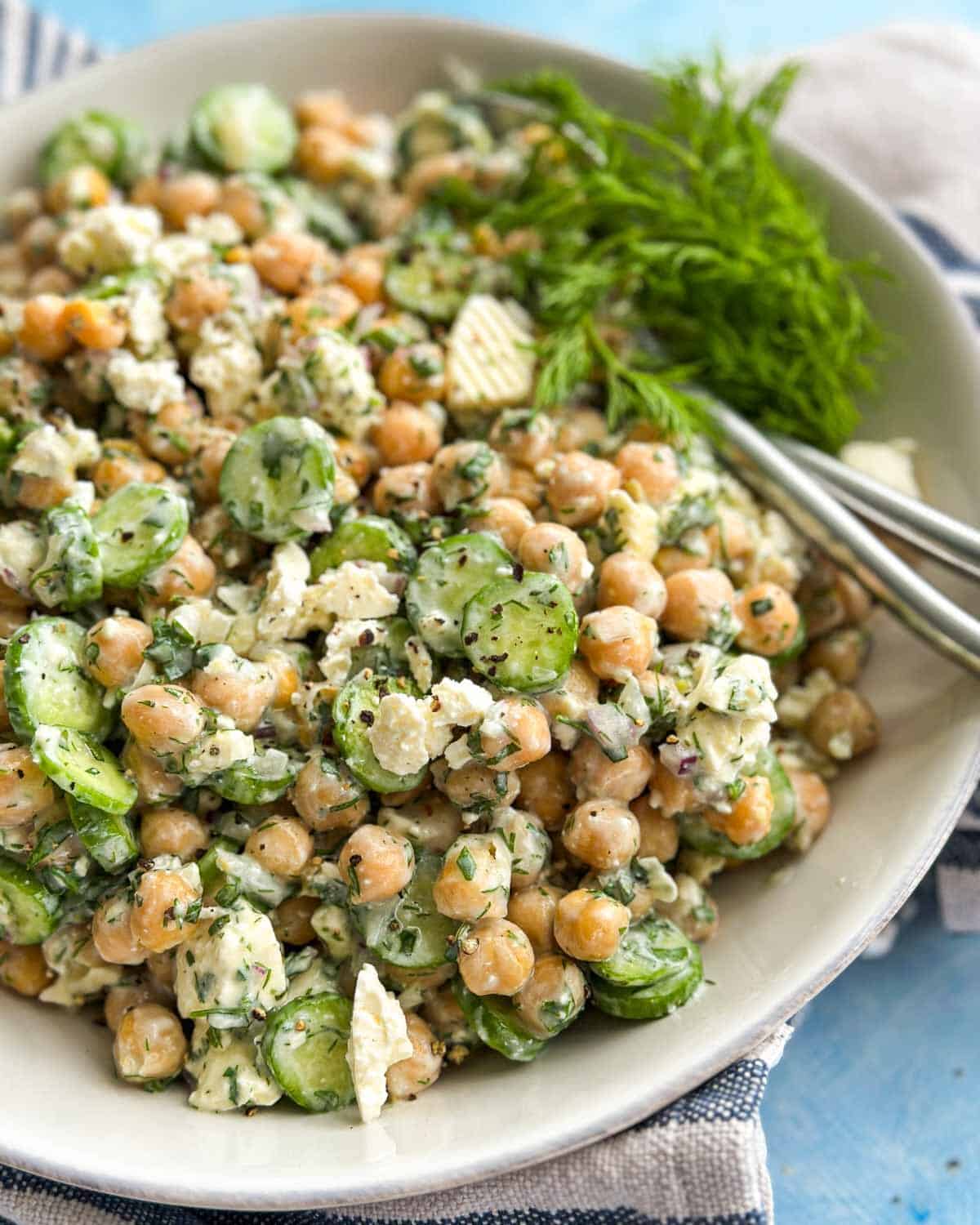 Close-up of creamy chickpea and feta salad with sliced cucumbers, fresh herbs, crumbled feta, and black pepper.