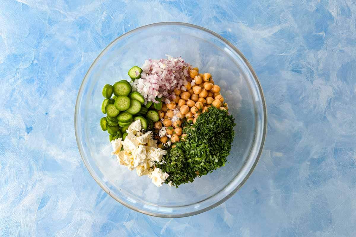 Chickpeas, cucumber, feta, red onion, and fresh herbs added to a glass bowl before mixing.