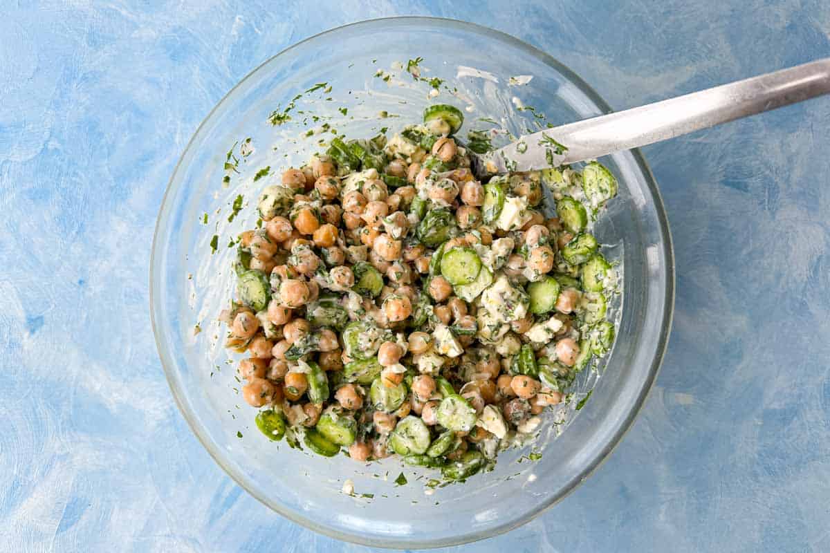 Creamy chickpea and feta salad mixed with cucumber, fresh herbs, and dressing in a glass bowl.
