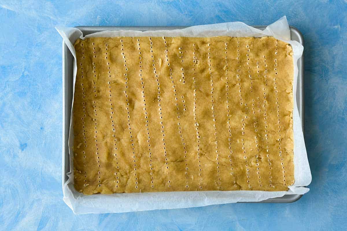 Berry crumble bar base pressed evenly into a parchment-lined baking tray and docked with a fork.