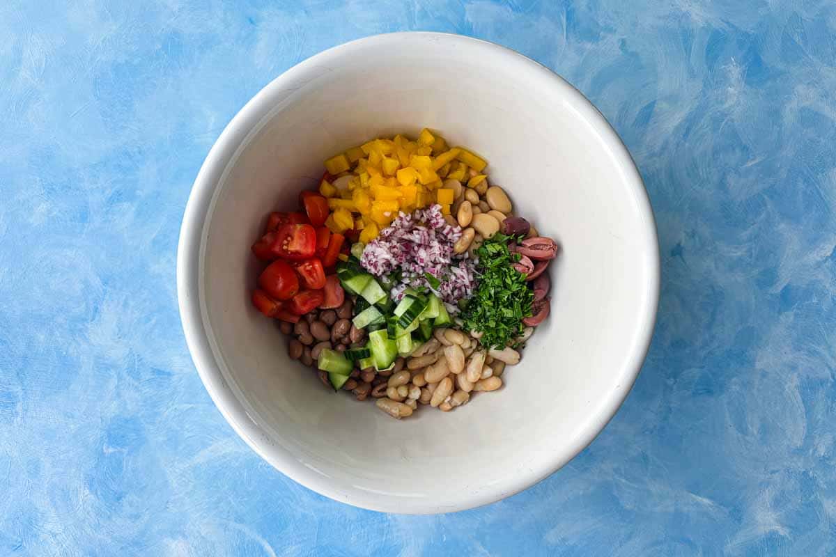 Chopped tomatoes, cucumber, bell pepper, olives, red onion, and parsley added to mixed beans in a bowl.