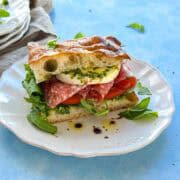 Focaccia bread sandwich filled with mozzarella, salami, tomatoes and arugula, served on a white plate with pesto.