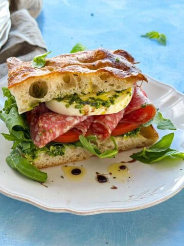 Focaccia bread sandwich filled with mozzarella, salami, tomatoes and arugula, served on a white plate with pesto.