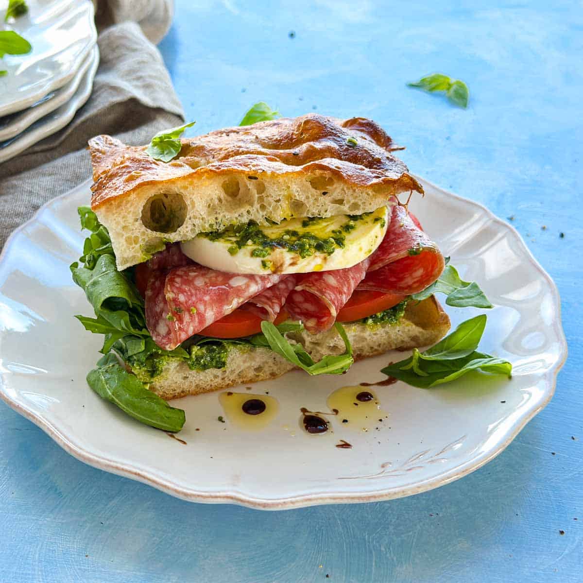 Focaccia bread sandwich filled with mozzarella, salami, tomatoes and arugula, served on a white plate with pesto.