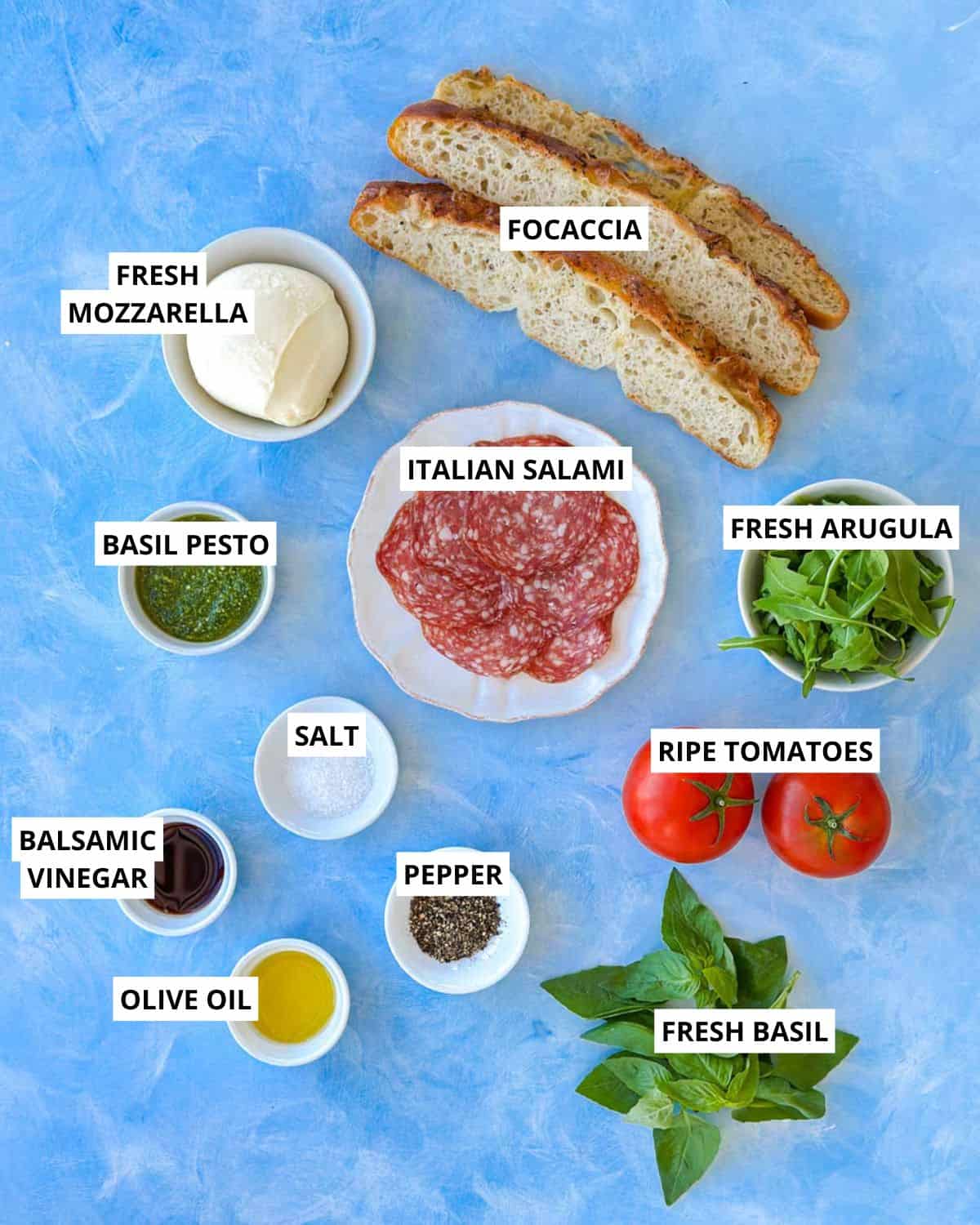 Ingredients for a focaccia bread sandwich including focaccia, fresh mozzarella, Italian salami, basil pesto, arugula, tomatoes and basil.