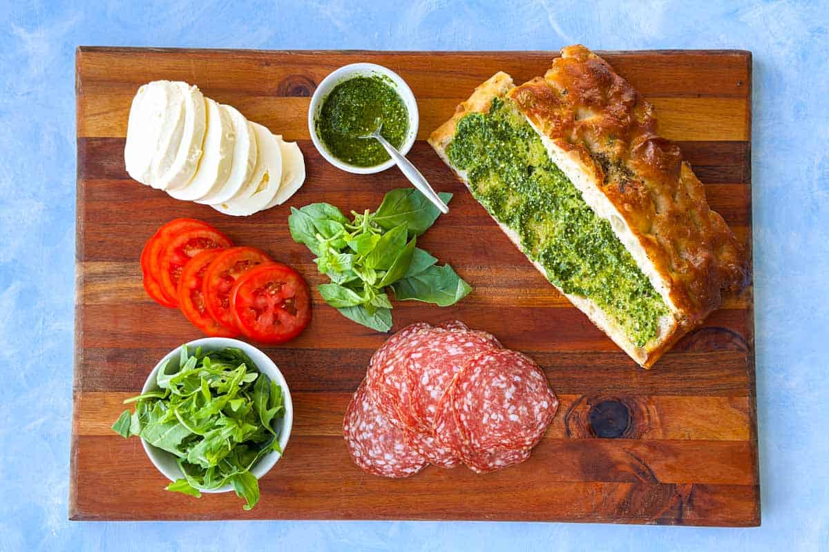 Focaccia bread sandwich ingredients arranged on a wooden board with pesto-coated focaccia, mozzarella, tomatoes, salami and arugula.